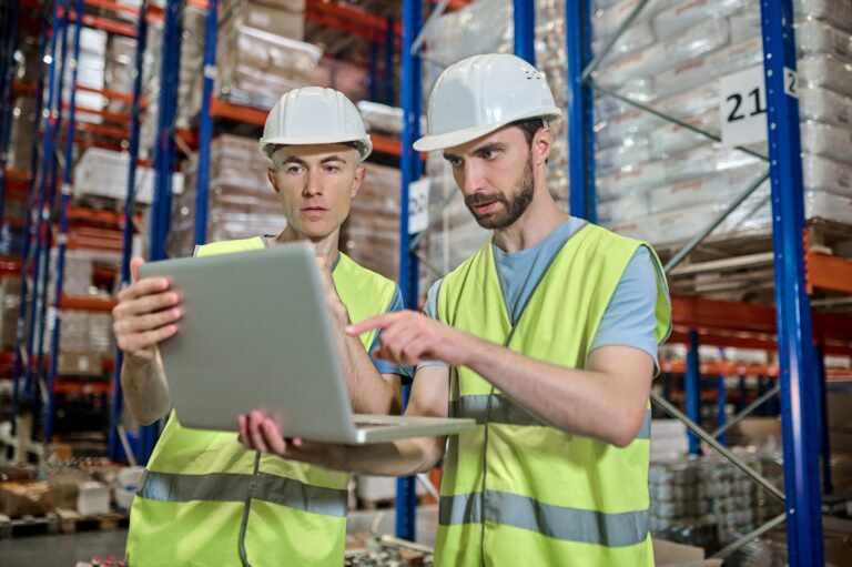 Dois homens de capacete em uma fábrica olhando um laptop